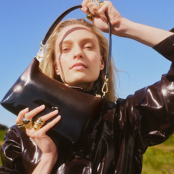 A women in a white shirt and cream pants has a black Saben Handbag across her body. The Saben handbag features a gold chain and gold saben lettering embossed at the top