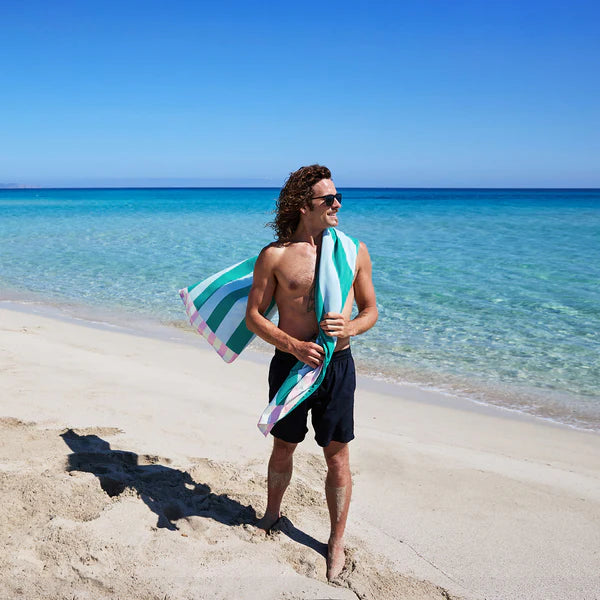 Man holding a striped towel on a beach with clear blue water and sky.