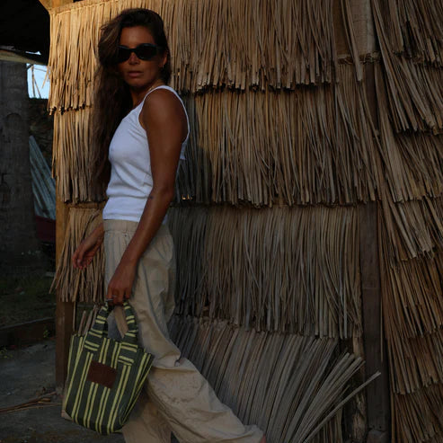 Woman standing in front of a thatched wall holding a striped bag.