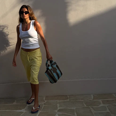 Woman in white tank top and yellow shorts standing against a white wall with a shadow, holding a black and green bag.