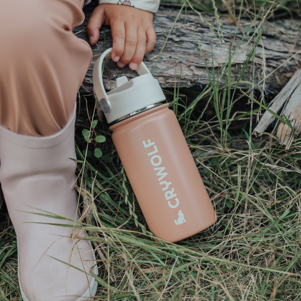 Lower portion of a child holding a Light orange colour sippy water-bottle with the text "crywolf" in white text with a wolf outline and a white coloured lid sitting on a lap