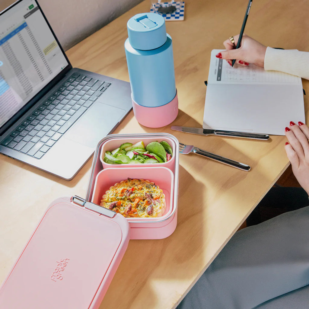 Pink bento box with salad and rice on a desk with a laptop and water bottle.