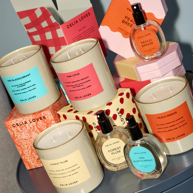 Collection of Celia Loves candles and perfume bottles on a gray surface with colorful boxes in the background.