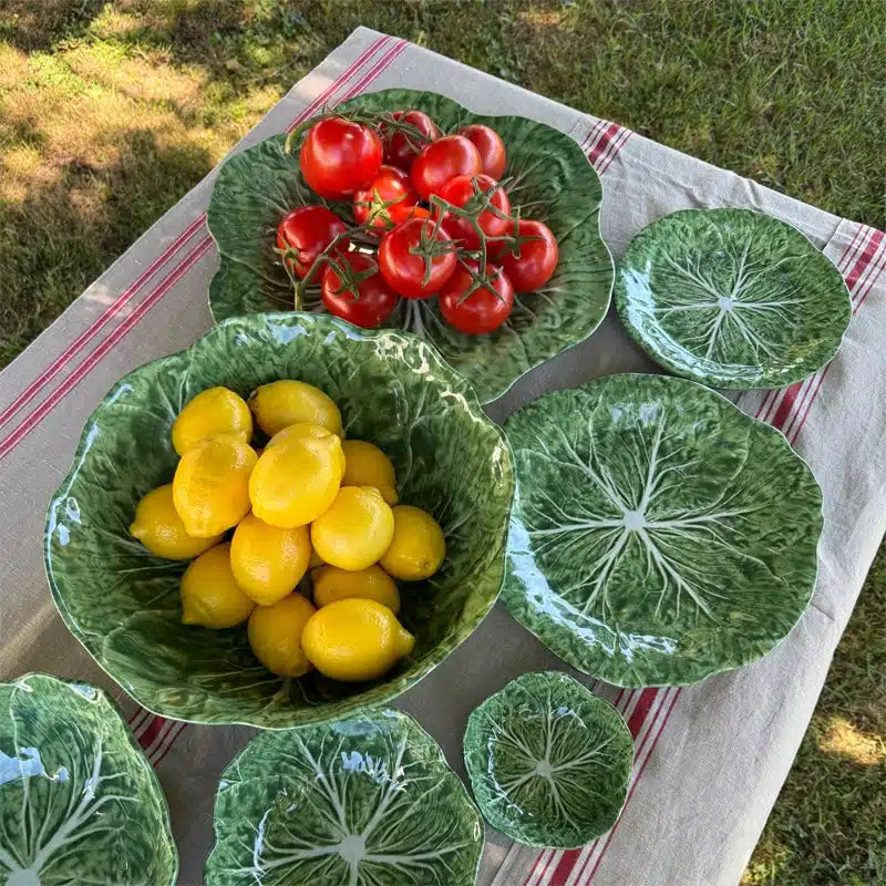 Green melamine plates with leaf patterns containing yellow fruits and red tomatoes on a checkered cloth outdoors.