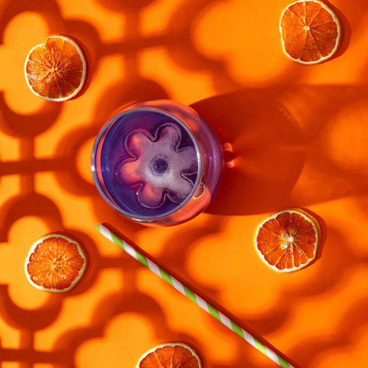 Purple drink with a flower-shaped ice cube in a clear glass, surrounded by dried orange slices on an orange patterned background.