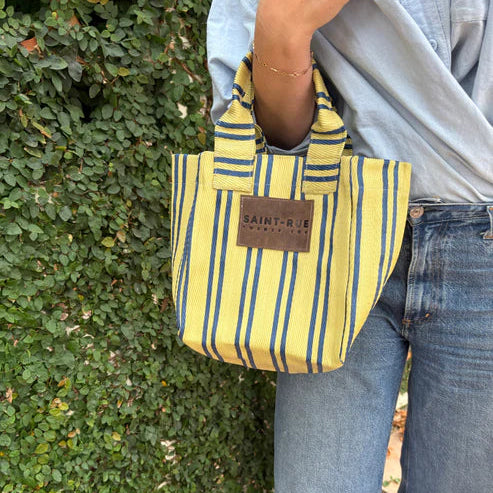 Person holding a yellow and blue striped bag with 'Saint-Rene' branding against a green hedge.