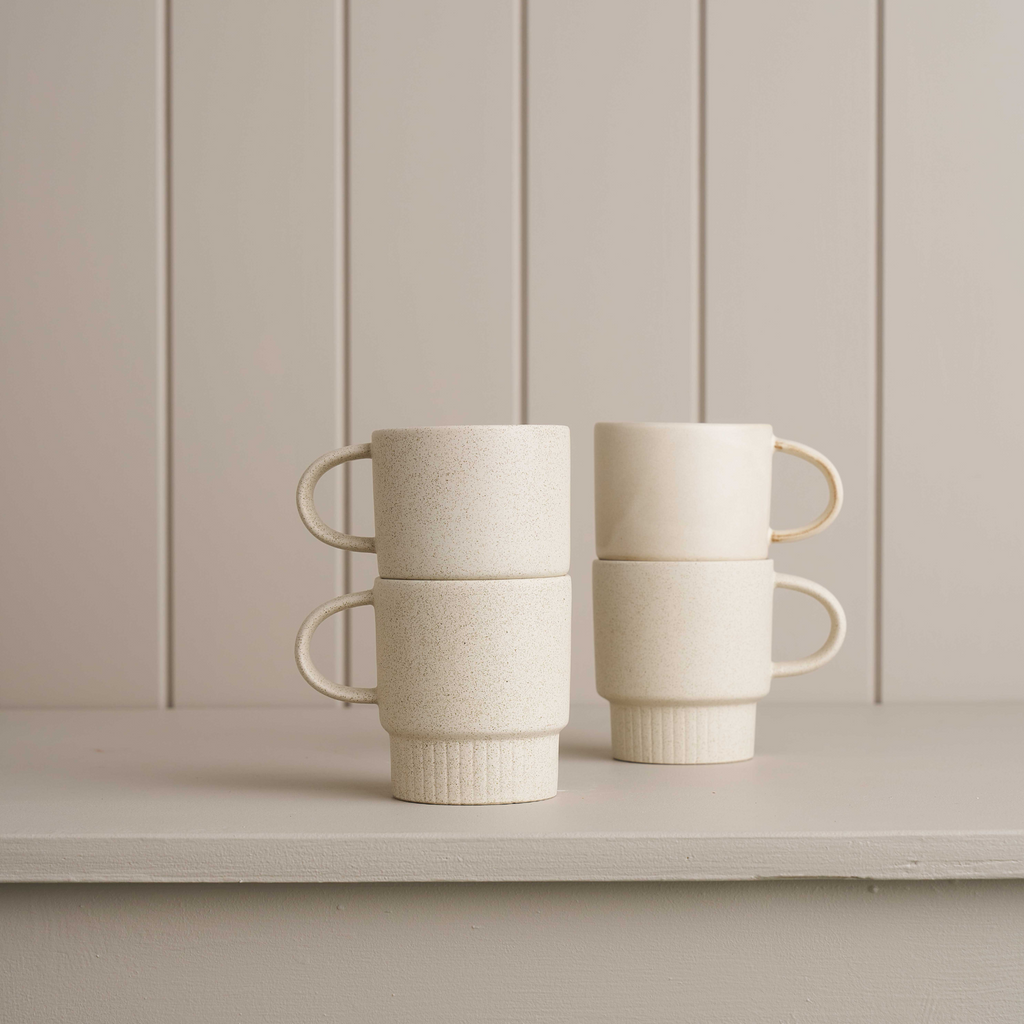 Two sets of a stack of two white ceramic mugs with a handle and narrow ribbed bottom on a grey mantle piece with a grey board wall behind it.