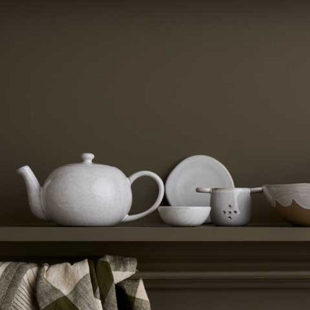A set of matching white ceramics including a tea strainer, coasters, mug, pinch pot and a tea pot on a green shelf against a green wall. 