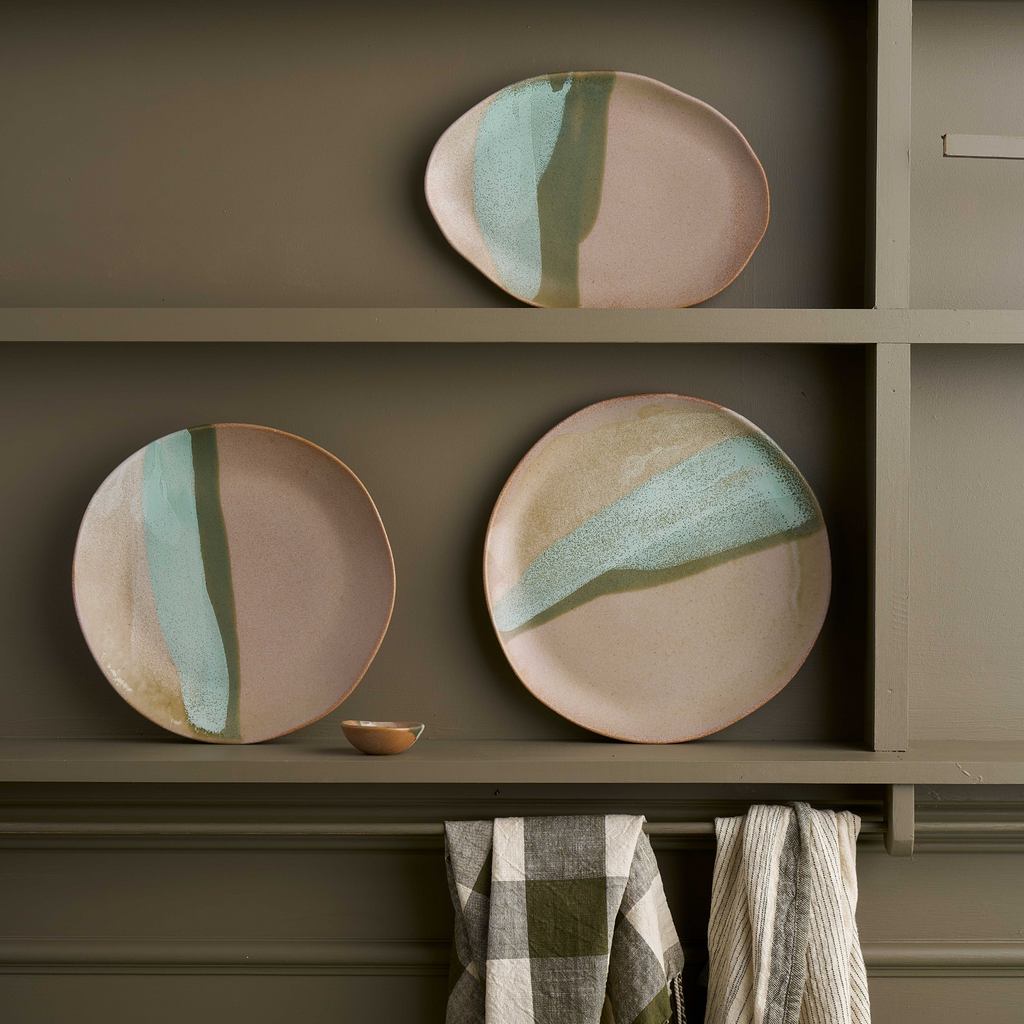Three various grey natural shaped ceramic serving trays with green and blue paint stroke features displayed on the shelves of a green shelving unit with some tea towels hanging off.