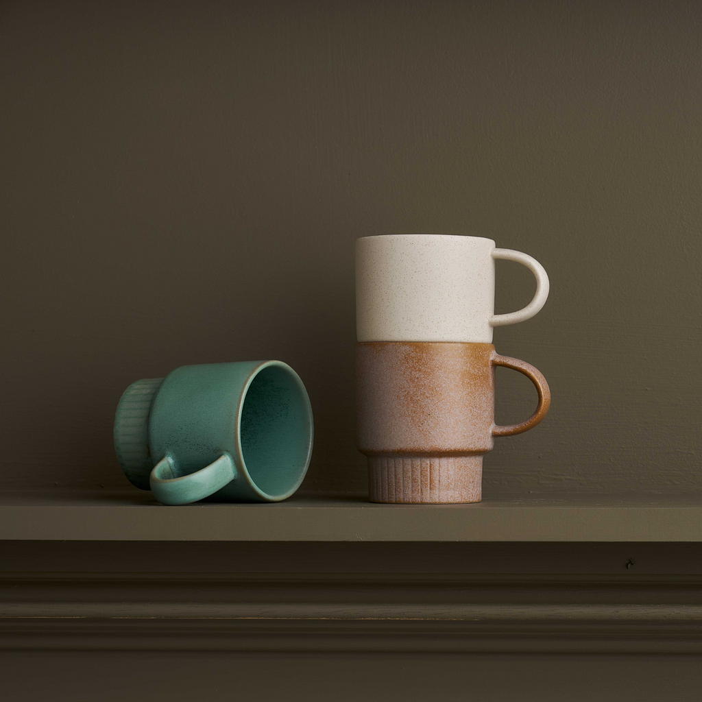  A stack of two ceramic mugs, one being white and another brown with a handle and narrow ribbed bottom and a blue mug rested on its side on a green mantle piece with a green wall behind it.