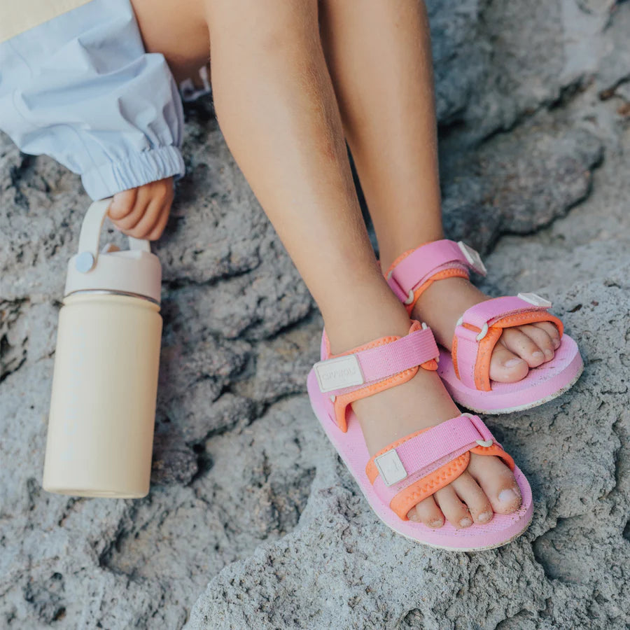 Child's feet wearing pink sandals on a sandy surface with a beige water bottle.