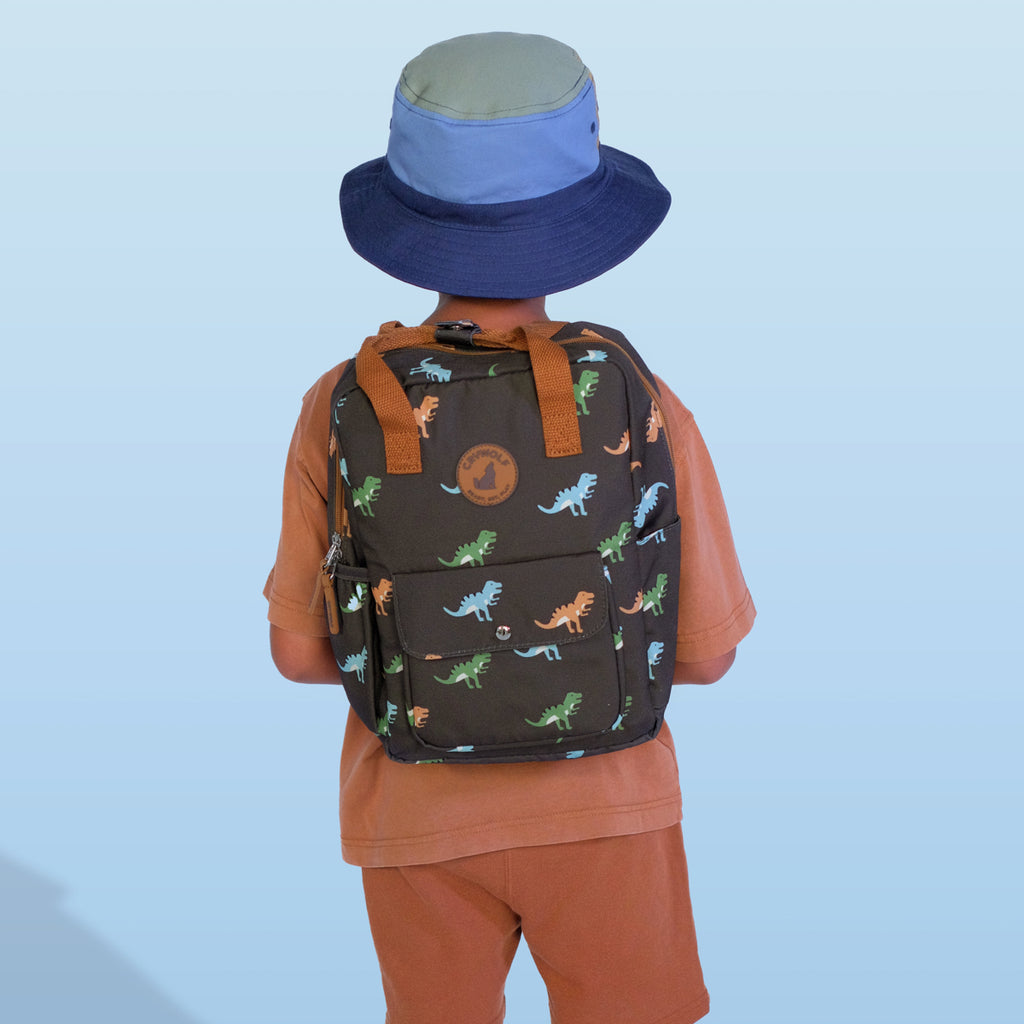 Child wearing a dinosaur-patterned backpack and blue hat on a light blue background