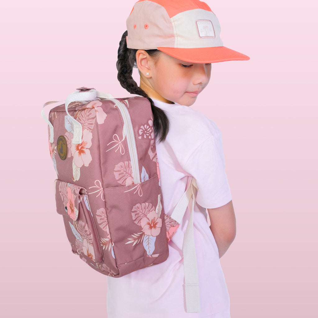 Child wearing a floral backpack and pink cap on a pink background