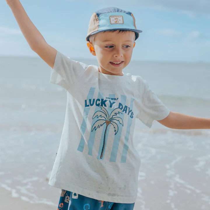 Child wearing a 'Lucky Days' t-shirt with a palm tree design on a beach.