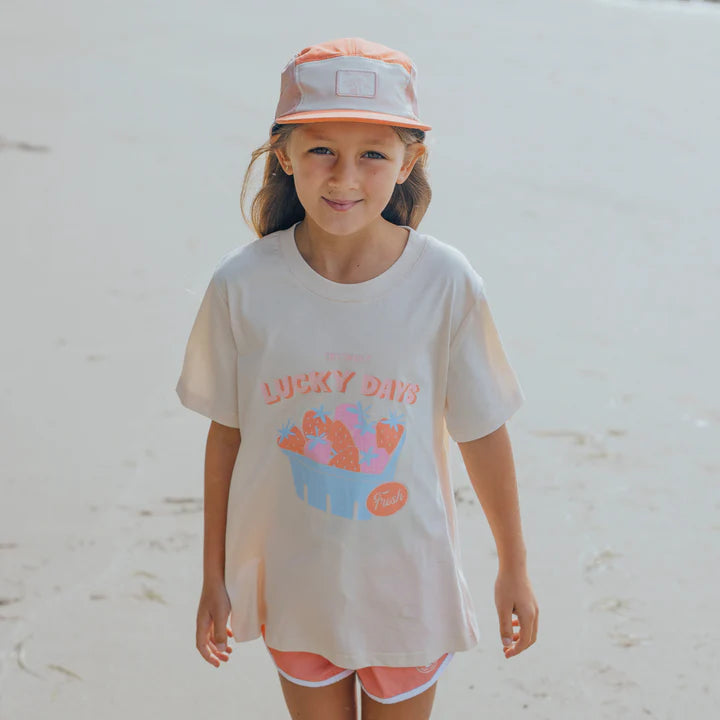 Young girl wearing a 'Lucky Days' t-shirt and cap on a sandy background