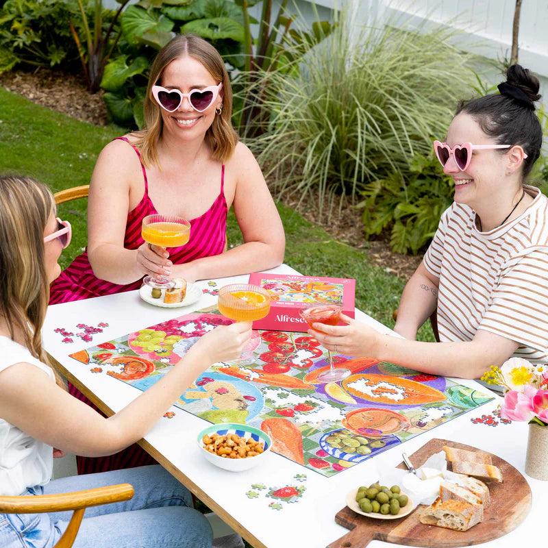 A group of people sitting around an outdoor  table drinking cocktails with a puzzle on the table