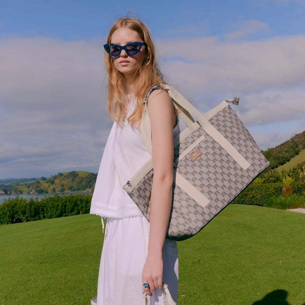 Woman in a white dress holding a patterned bag outdoors with greenery and blue sky in the background