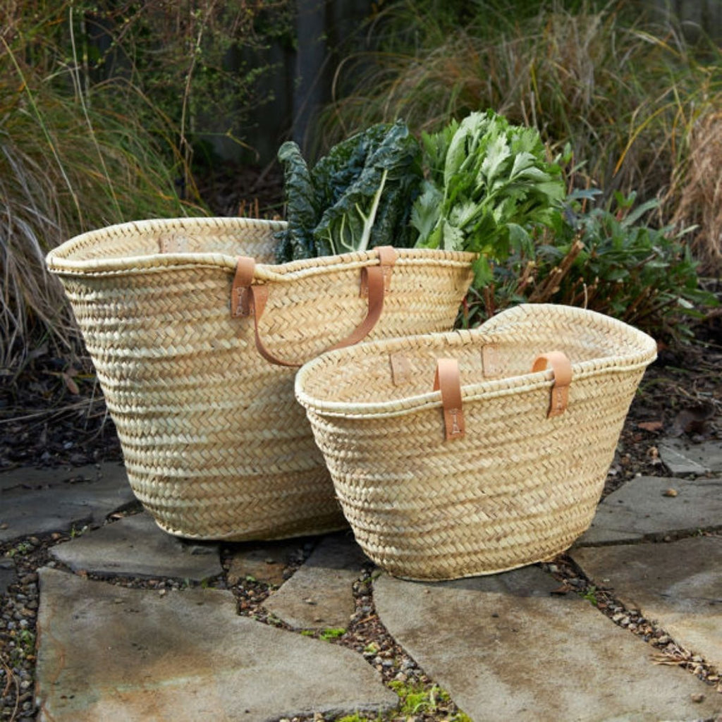 Two woven baskets with handles on a stone path with plants in the background