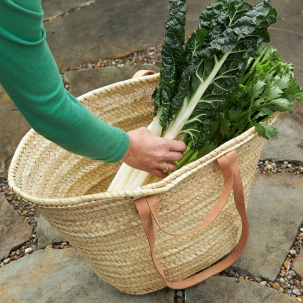 Person holding a woven basket with green leafy vegetables on a stone surface