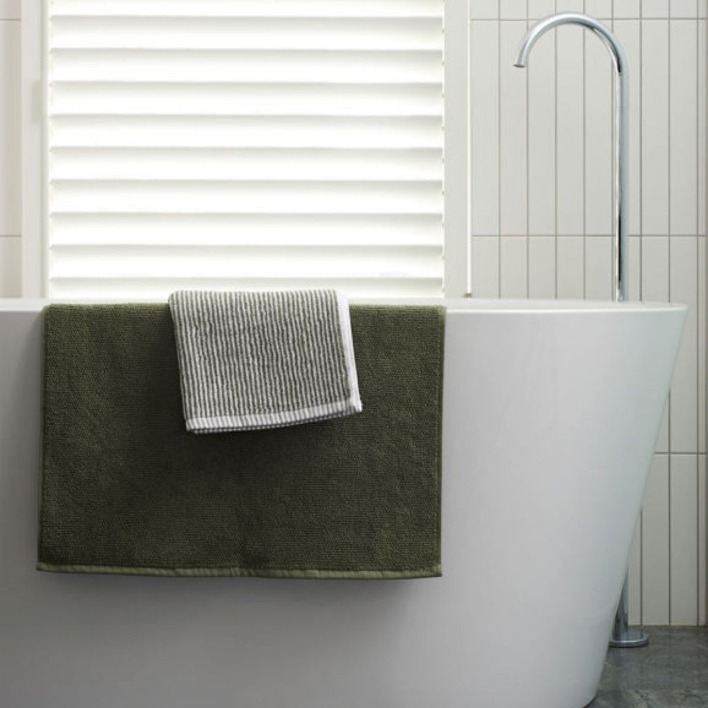 Green bath mat on a white bathtub with a towel draped over it in a bathroom setting.
