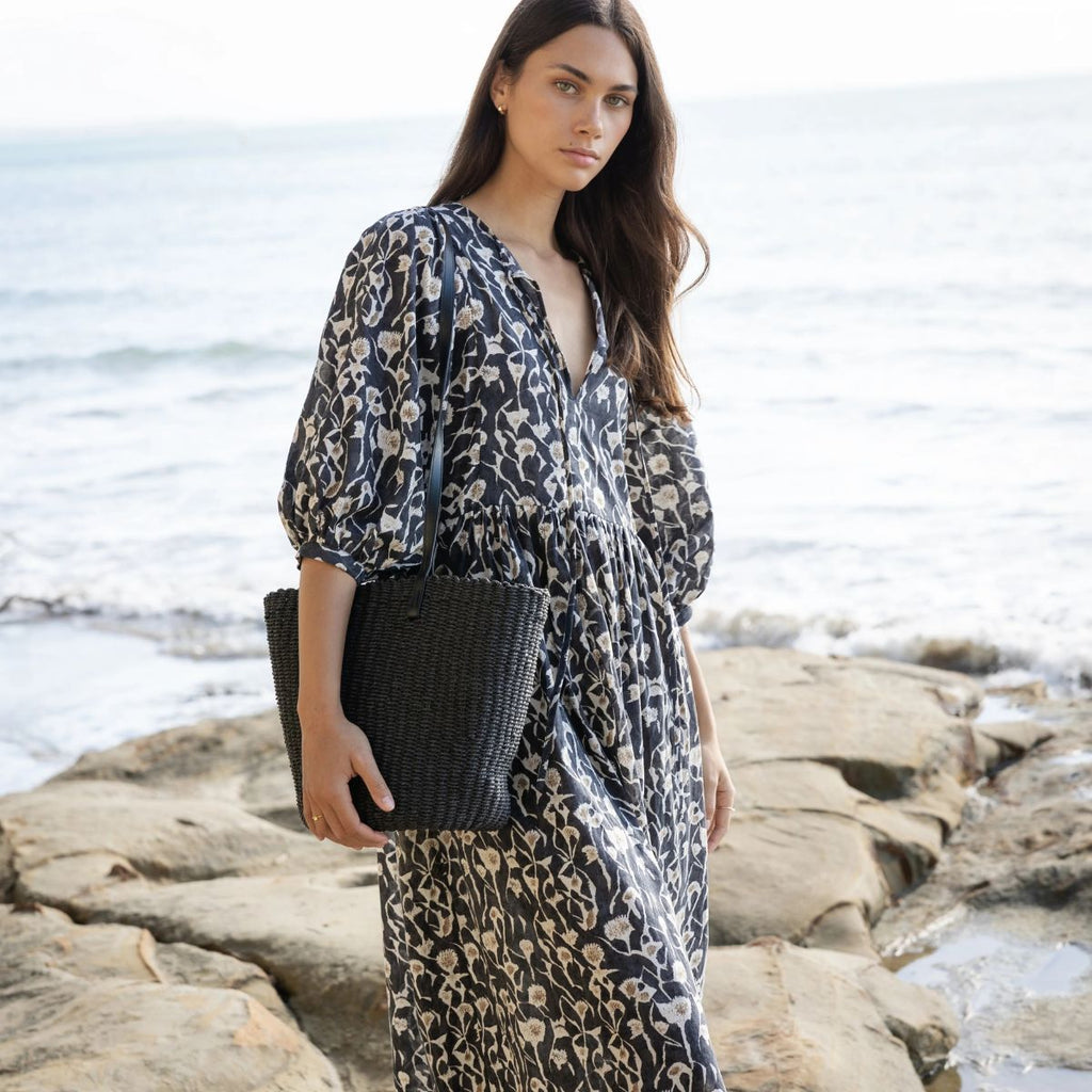 Woman in a floral dress standing on rocks by the ocean, holding a black woven basket