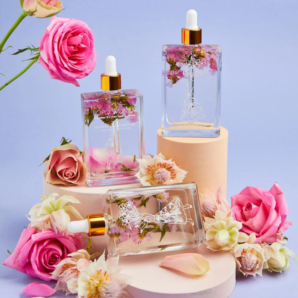 Three glass perfume bottles with floral designs on a purple background with pink and white flowers.