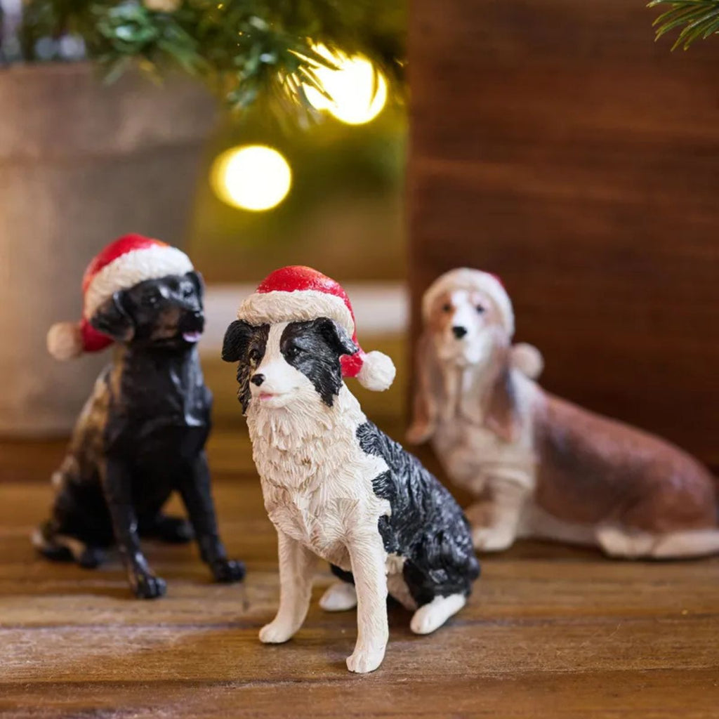 Three dog figurines wearing Santa hats on a wooden surface with a blurred Christmas tree in the background.