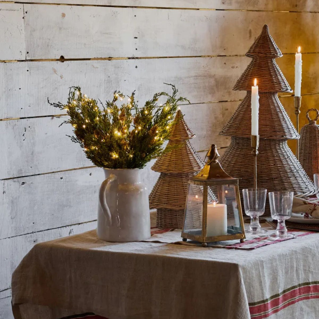 Decorative table setting with wicker Christmas trees, candles, and a vase of lights against a wooden wall.