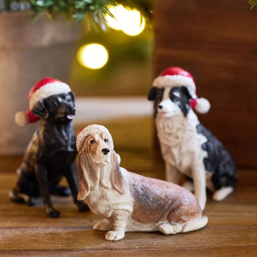 Three dog figurines wearing Santa hats on a wooden surface with a blurred festive background.