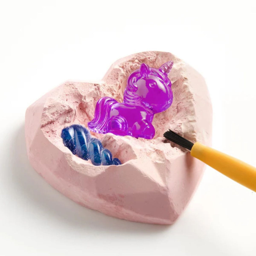 Pink heart-shaped mold with a purple unicorn and blue jelly-like substance, being filled with a yellow tool.