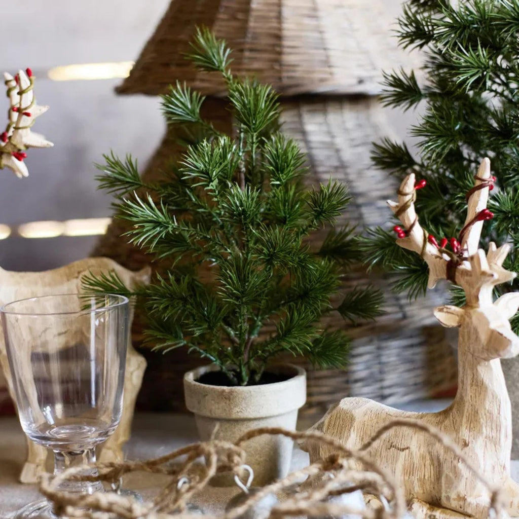 Decorative setting with a small potted plant, glass, and wooden reindeer with berries on a blurred background.