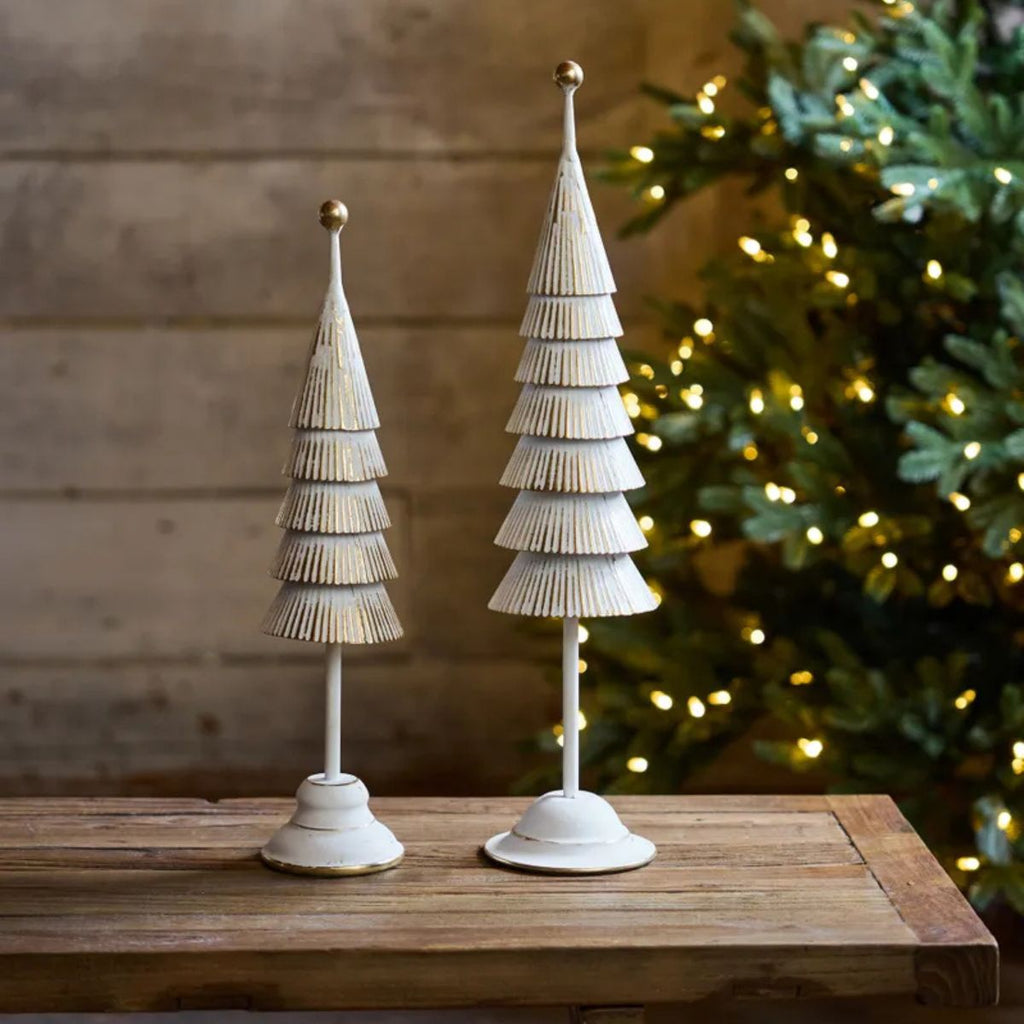 Two decorative Christmas trees on a wooden surface with a blurred Christmas tree in the background.