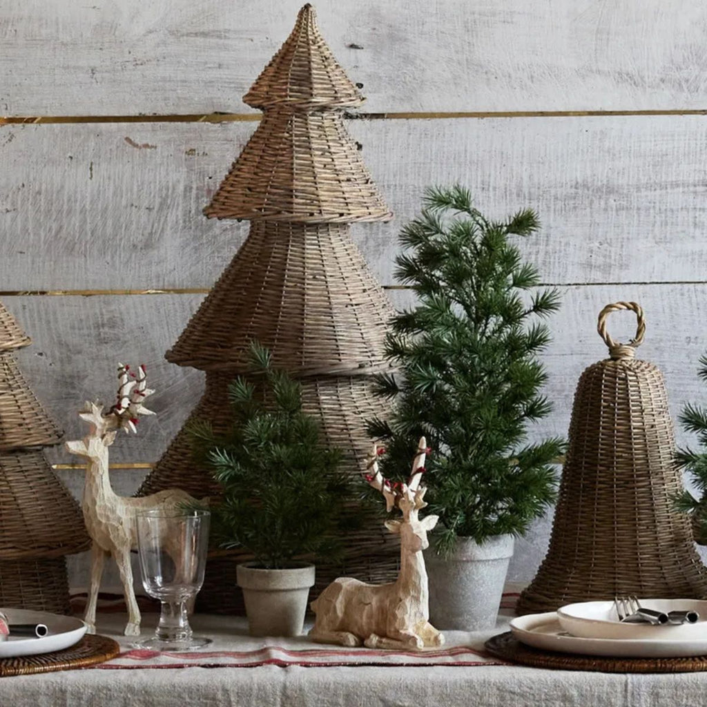 Decorative table setting with wicker Christmas trees, small potted trees, and deer figurines against a wooden wall.