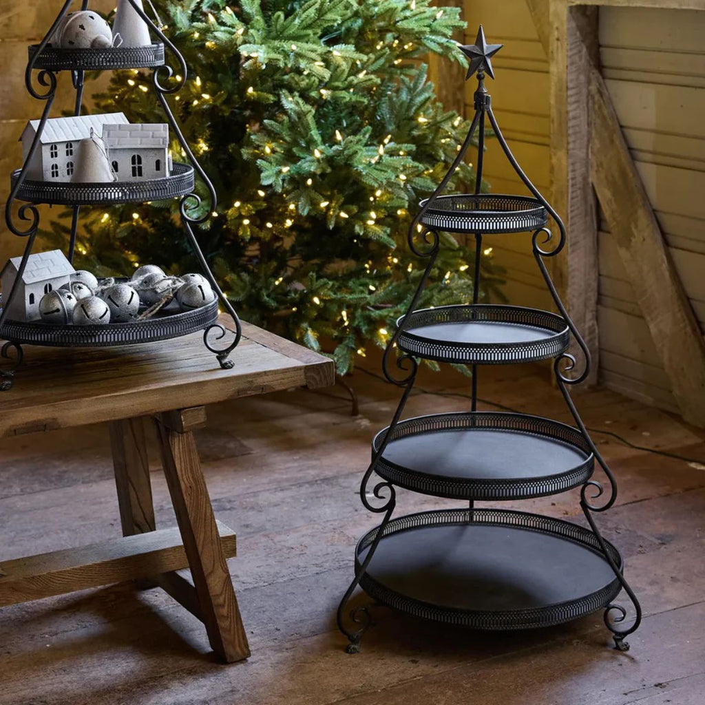 Decorative metal tiered stand with small houses and balls in front of a Christmas tree.