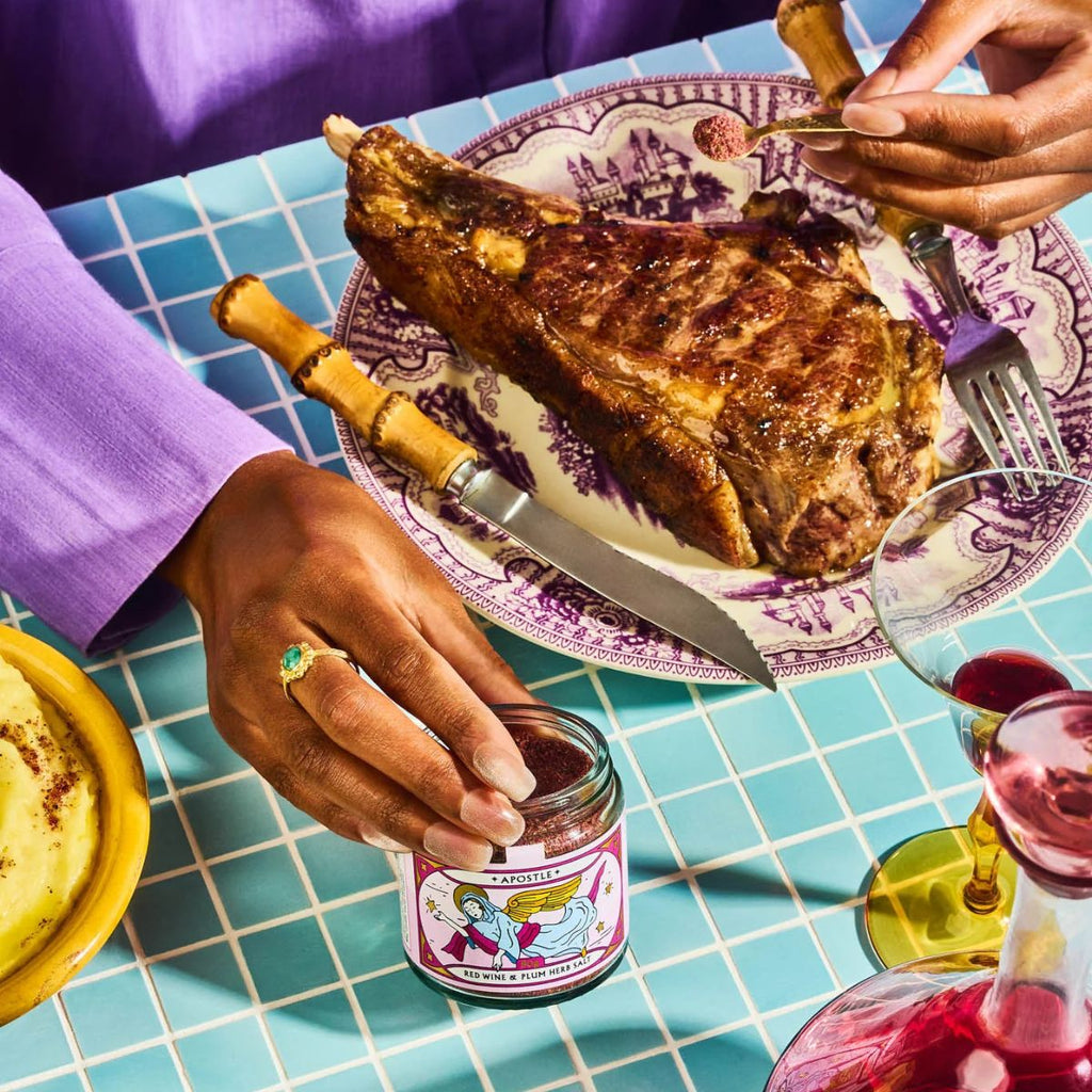 Person seasoning a steak with a jar of seasoning on a table with a steak and wine glass.
