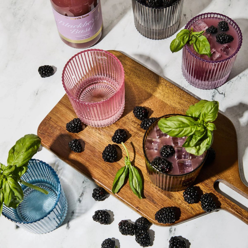 Pink glasses with blackberry cocktails on a wooden board, surrounded by blackberries and basil leaves.