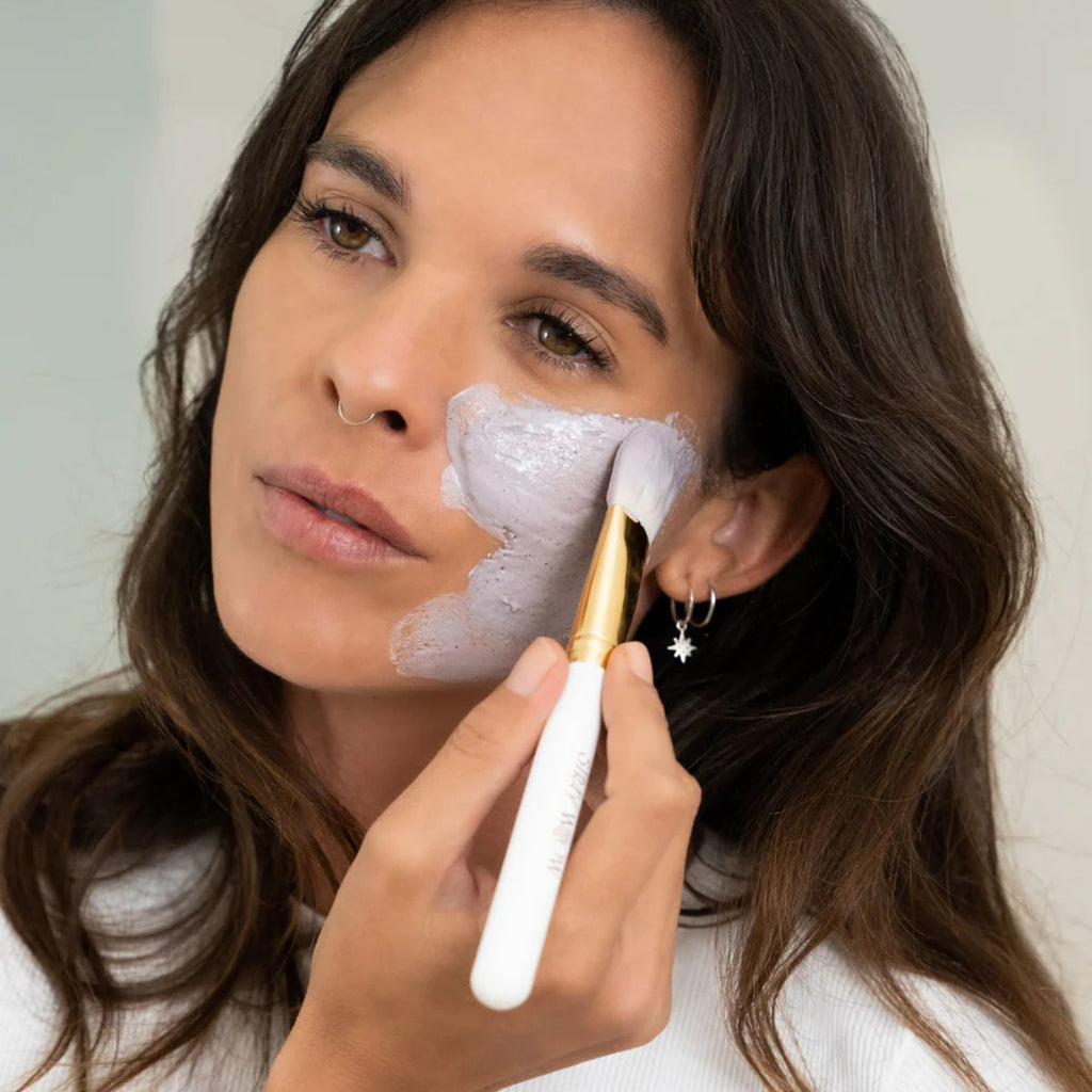 Woman applying a facial mask with a brush to her face.