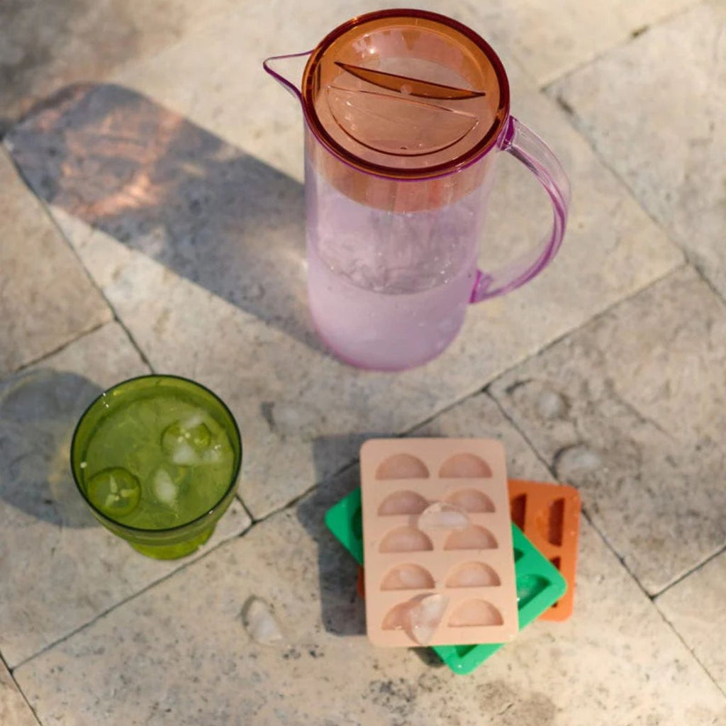 Pink pitcher, green cup with ice, and colorful ice cube trays on a stone surface.