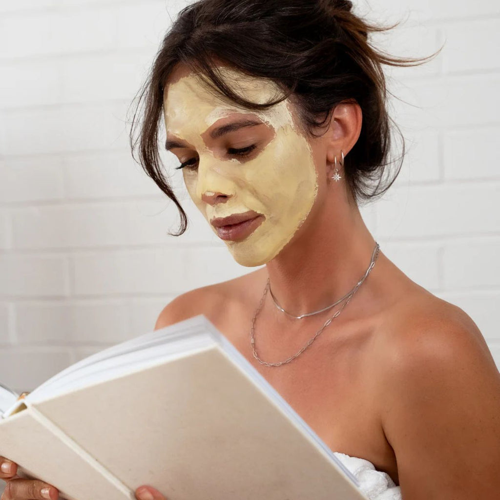 Woman applying facial mask and reading a book in a bathroom.