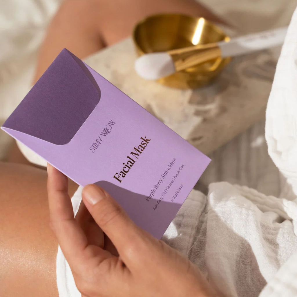 Hand holding a purple facial mask package with a blurred background of skincare products.