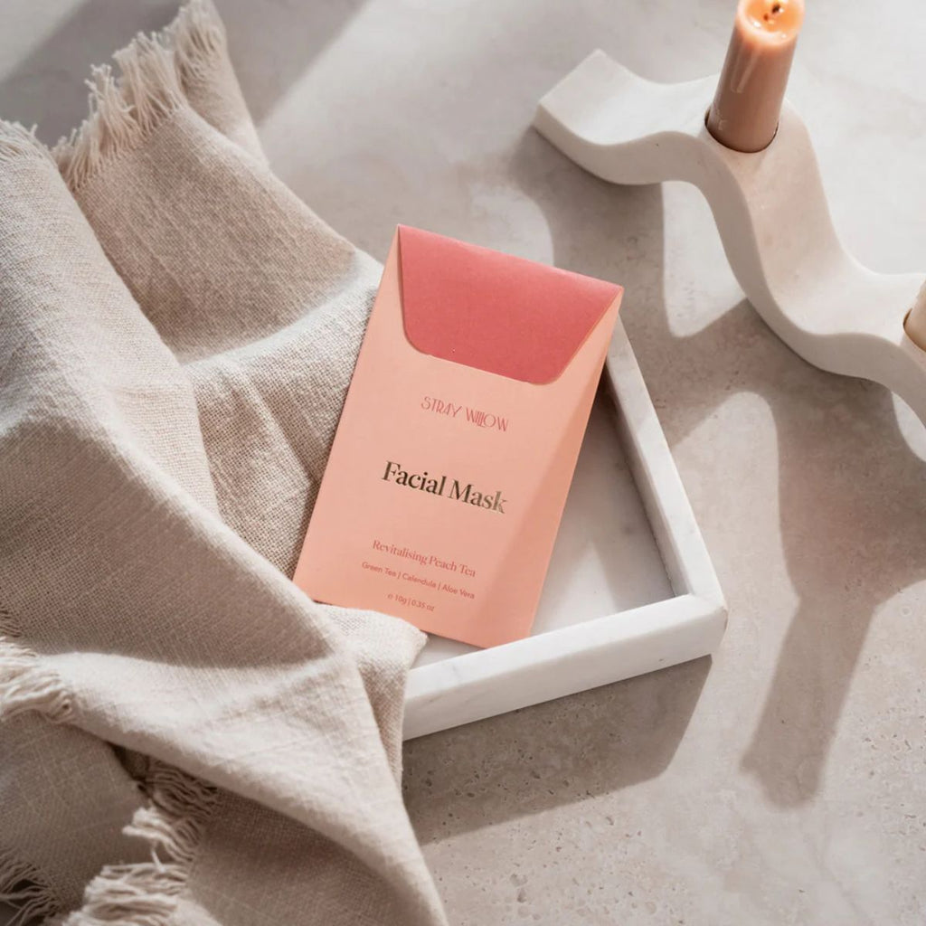 Pink facial mask packaging on a white tray with a beige towel and candle in the background