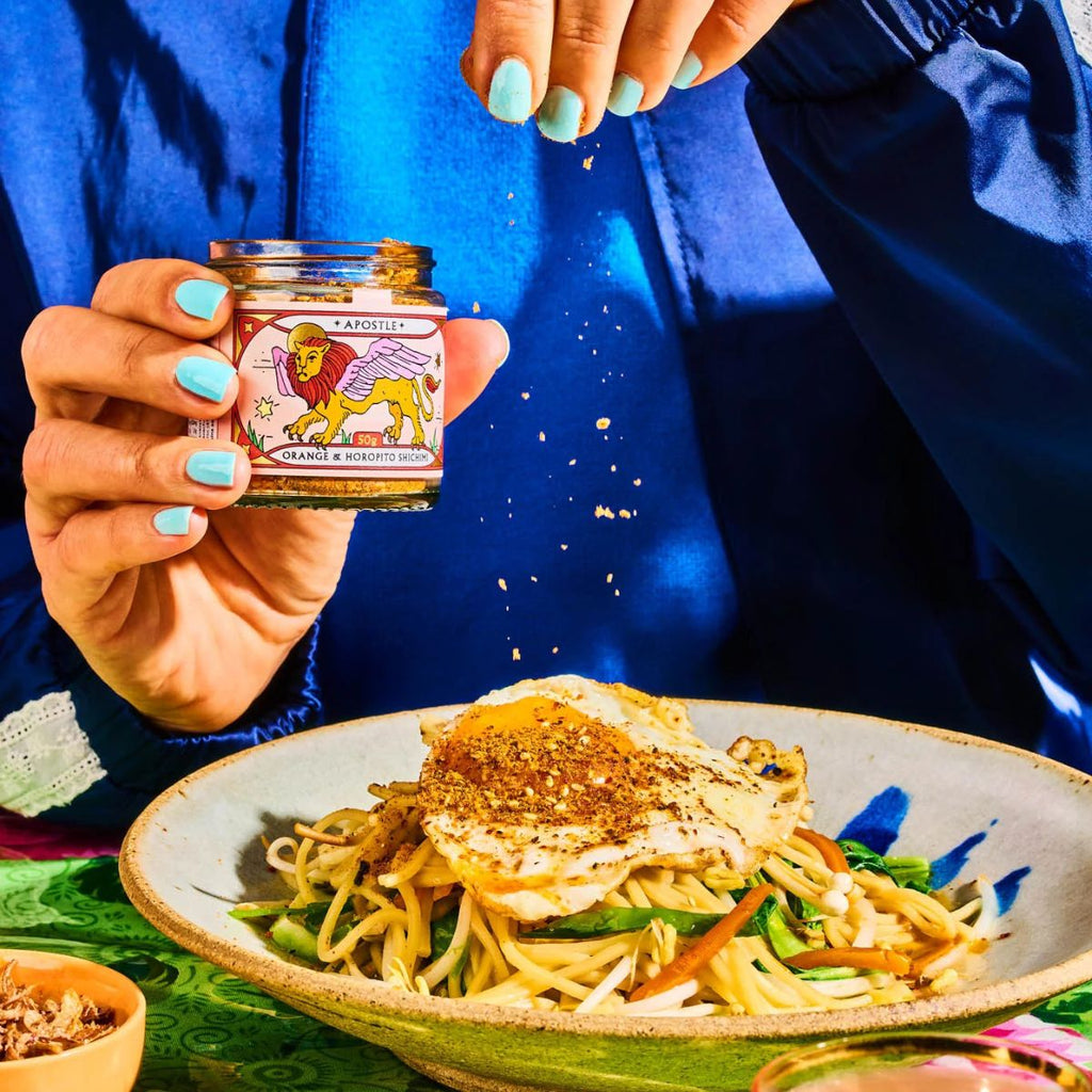 Person sprinkling seasoning from a jar onto a dish of noodles with vegetables.