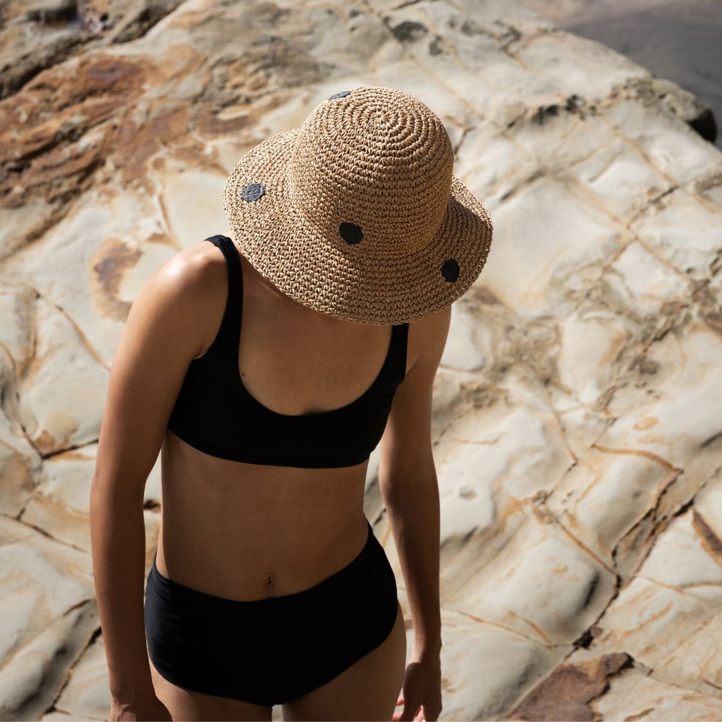 Person wearing a straw hat and black swimsuit against a stone wall.