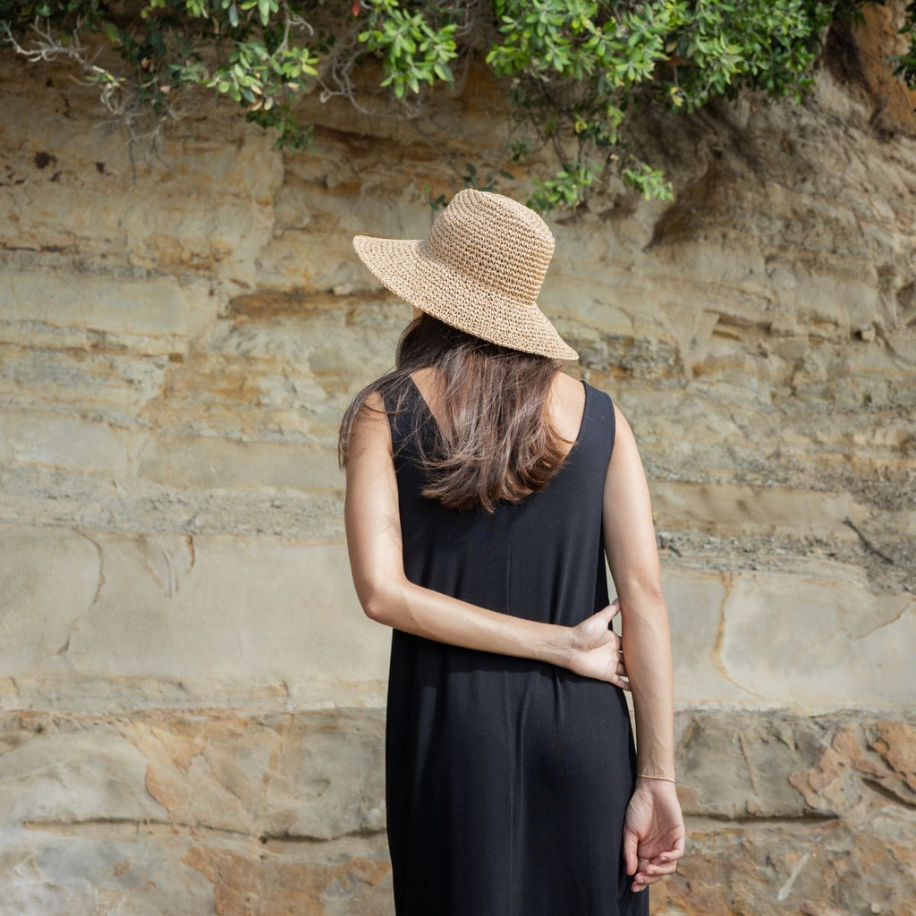 Woman wearing a black dress and straw hat against a stone wall with greenery.