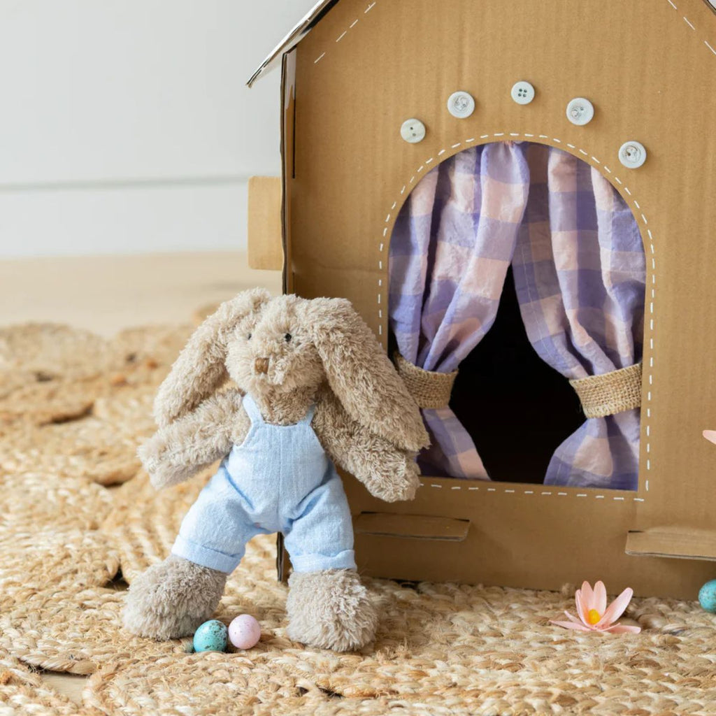 Cardboard house with a plush bunny toy and Easter eggs on a woven surface.