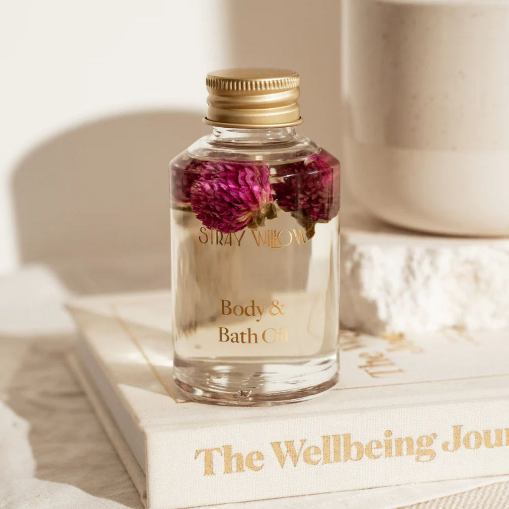 Glass bottle of 'Body & Bath Oil' with flowers on a book titled 'The Wellbeing Journey'.