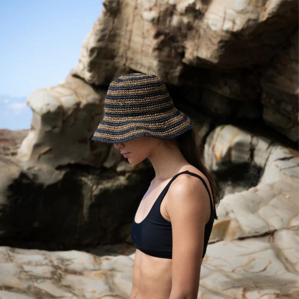 Woman wearing a black bikini and striped bucket hat standing against a rocky background