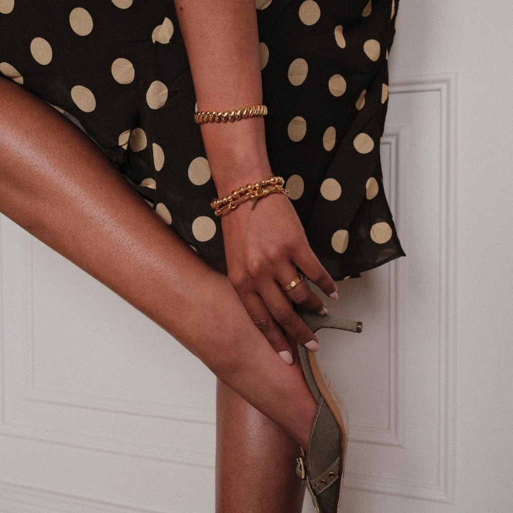 Person wearing a black dress with gold polka dots, holding a sandal, with a neutral background.