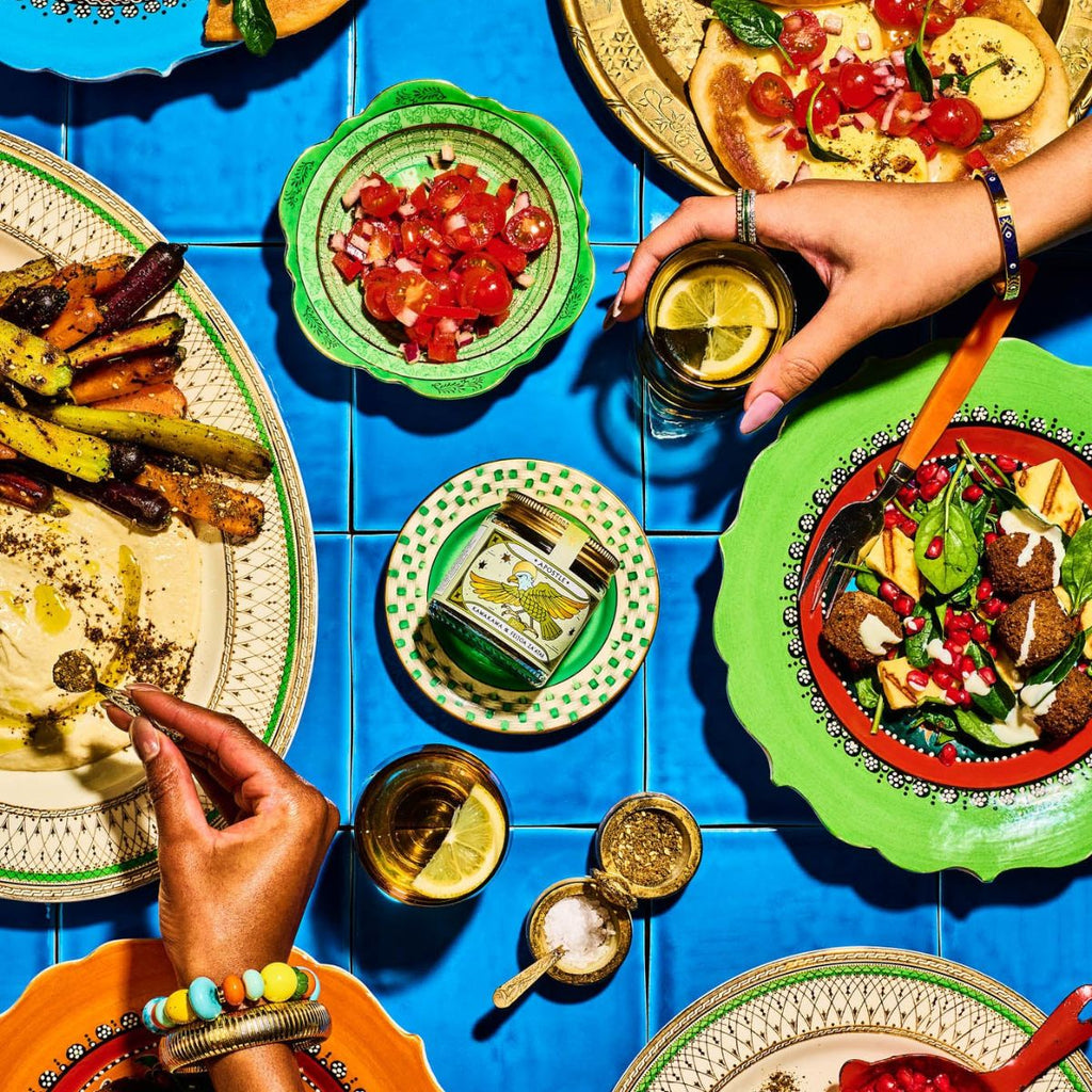 Colorful table setting with various dishes and hands interacting with food on a blue tiled surface.