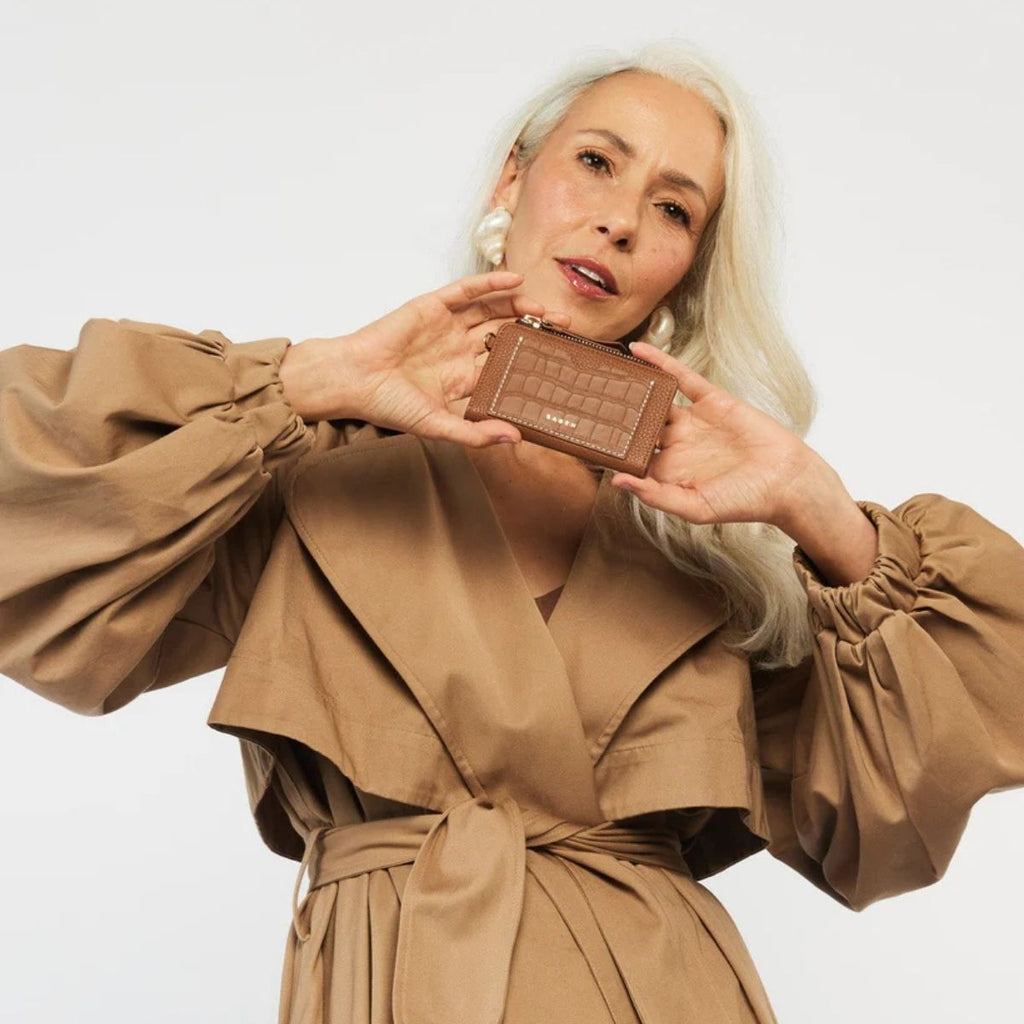 Woman in a beige trench coat holding a brown wallet against a light background